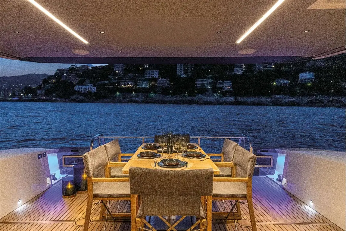 Dining table set with plates and glasses on a luxury yacht deck, with a sunset view of the coastline and water.